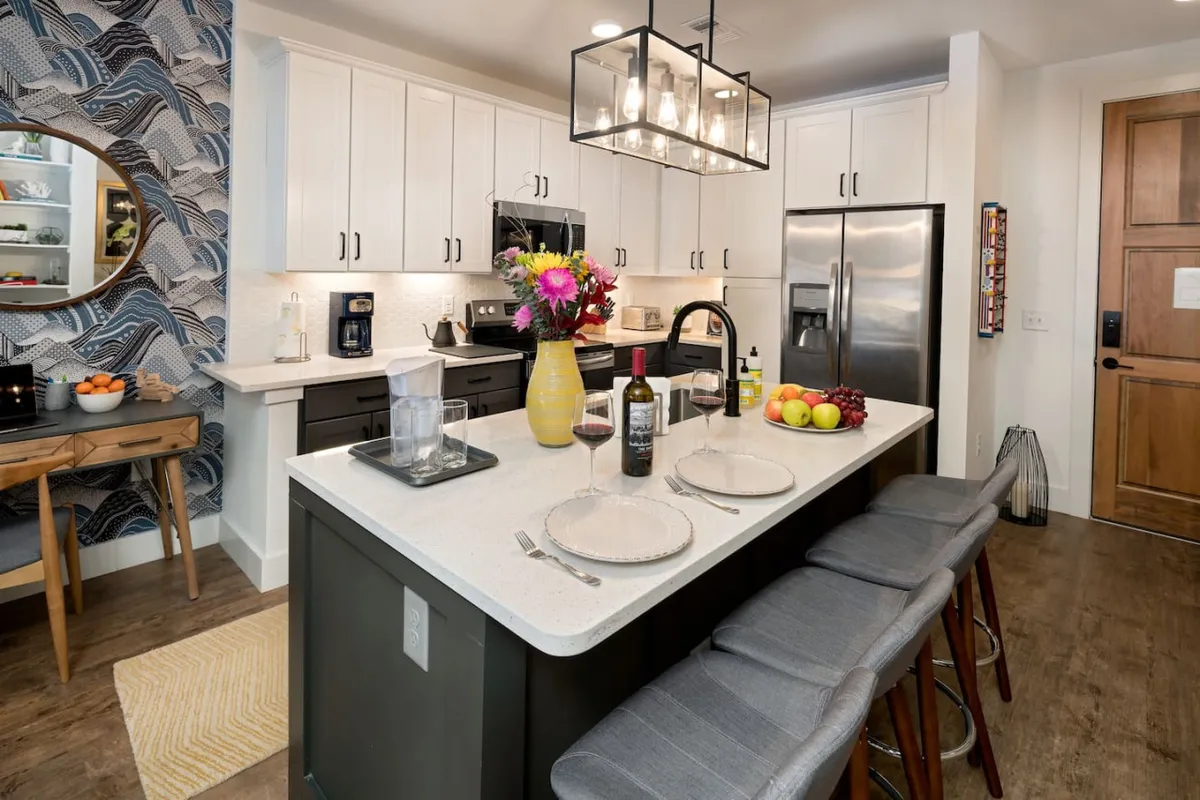 This image centers on the kitchen island, complete with a floral centerpiece and modern kitchen appliances visible in the background. Sleek bar stools line the island, inviting guests or residents to sit and enjoy meals. The space feels warm and welcoming, emphasizing both style and functionality.