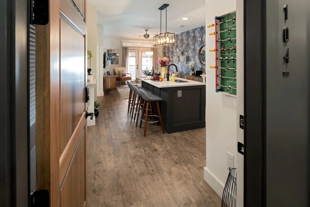 The kitchen island is visible adjacent to the front door, with bar stools and wall art adding visual interest. A foosball table is also present, suggesting a multi-functional, social space. The mood is cheerful and lively, ideal for gatherings and everyday living.