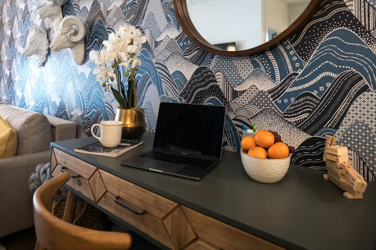 A contemporary workspace featuring a decorative wall with a wave pattern, a desk set up with a laptop and accessories, and a mirror reflecting light to create a bright, inviting atmosphere. The scene suggests a place ideal for productivity and enjoying a cup of coffee.