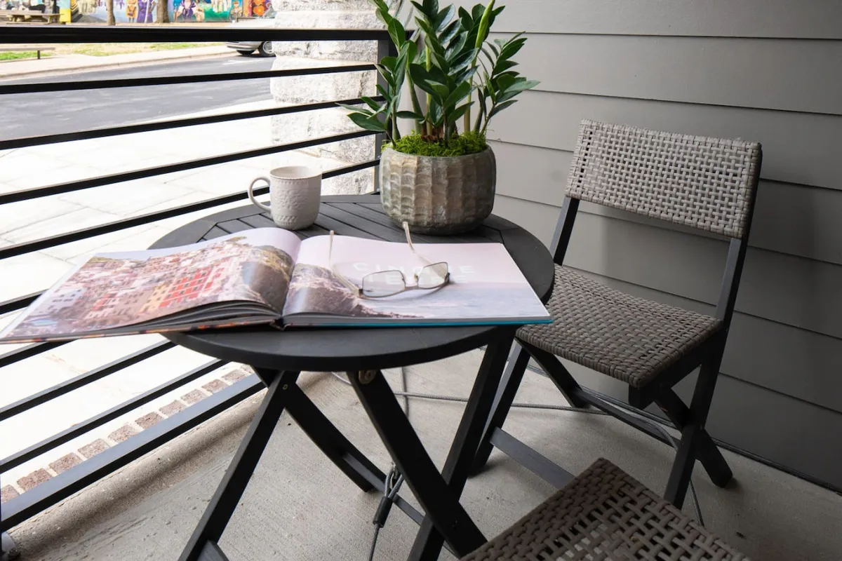 A cozy private patio is shown, featuring a small table with an open magazine, a ceramic mug, a potted plant, and a pair of glasses. The setting suggests a tranquil outdoor nook perfect for relaxing, reading, or enjoying a quiet drink while feeling connected to the urban surroundings.