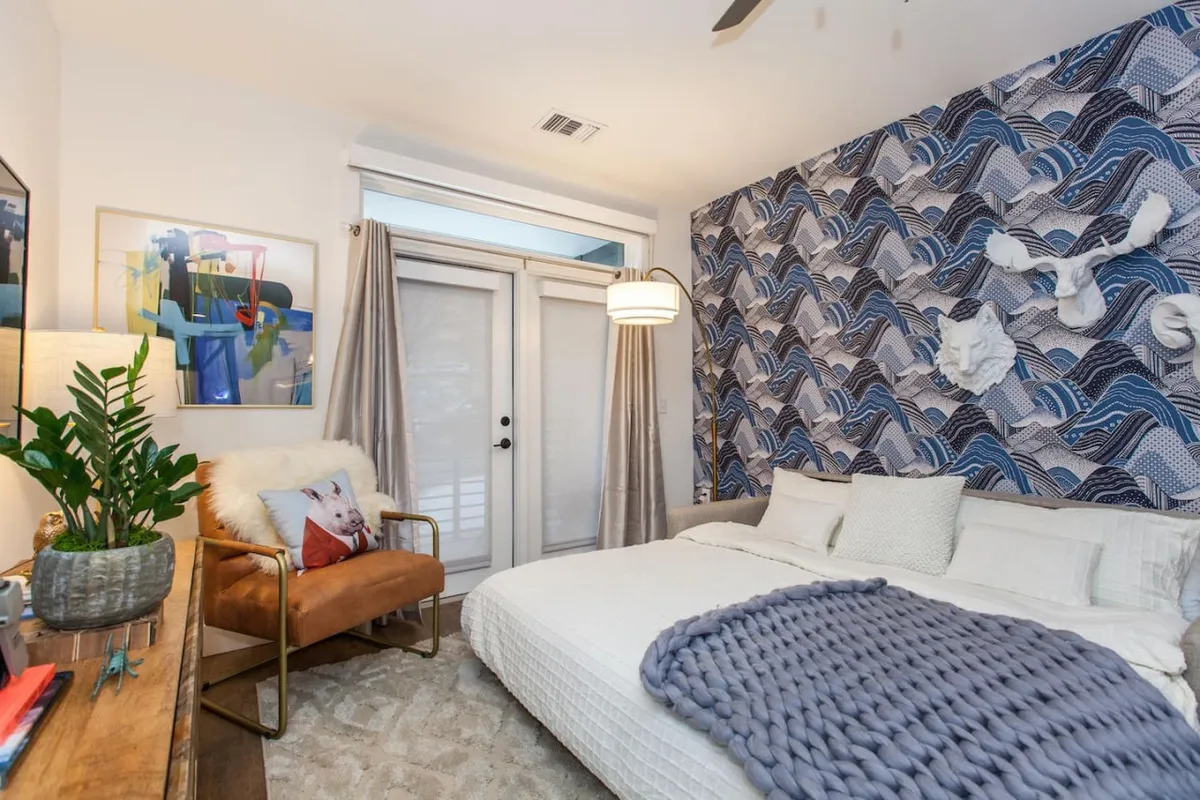 A tranquil bedroom corner featuring a cozy armchair beneath textured wave-patterned walls and a neatly made bed with a throw blanket. Soft natural light suggests relaxation, reading, or enjoying brunch by the window, reinforcing the retreat-like ambiance.