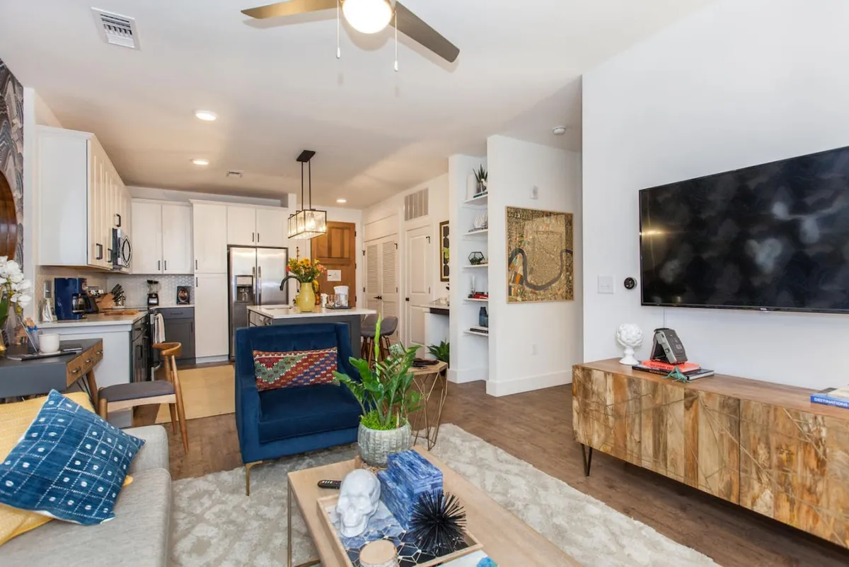 A bright, open living area featuring a blue armchair with decorative pillows, a wooden coffee table, a large TV mounted on the wall, and a glimpse of the kitchen area in the background. Unique wall art adds a touch of personality to the contemporary setting.