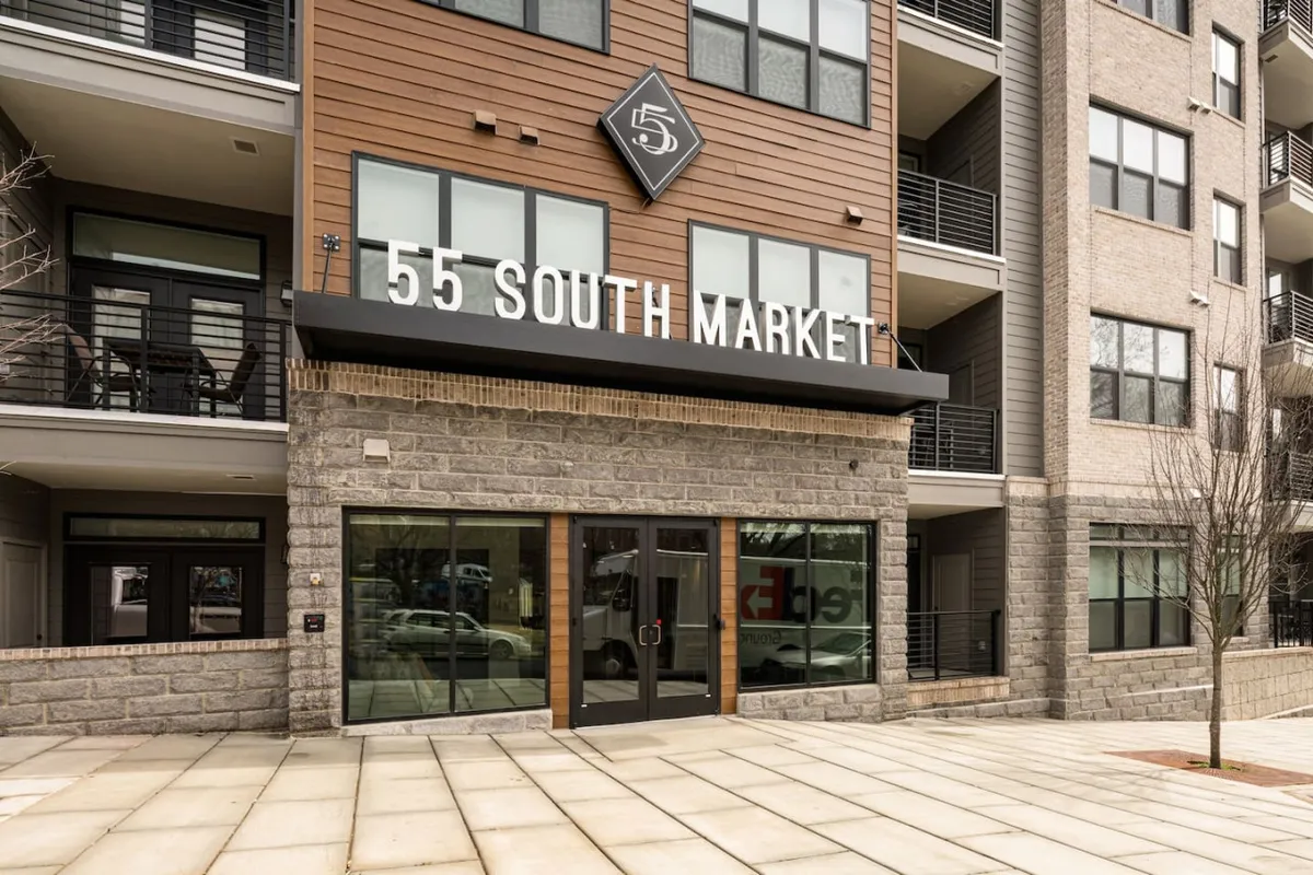 The exterior of a modern building is shown, with large windows and the visible signage of '55 South Market.' The entrance suggests easy access to urban life, while the suggestion of a patio area hints at communal or private outdoor spaces connected to the lively city environment.
