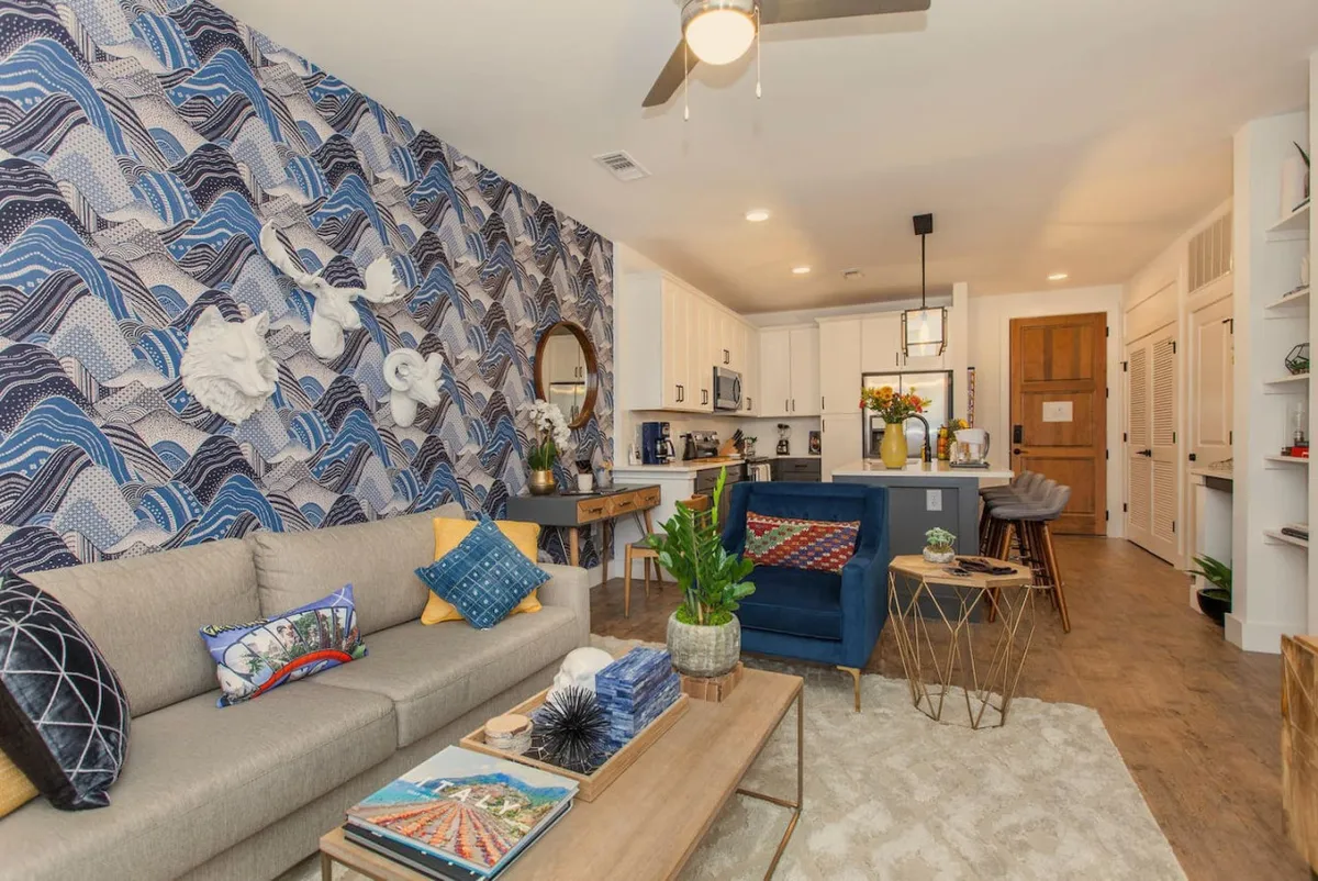 A comfortable seating area accented by textured wave-patterned wallpaper, colorful throw pillows, a stylish coffee table, and a green potted plant. The space is perfect for casual dining or relaxed conversation.