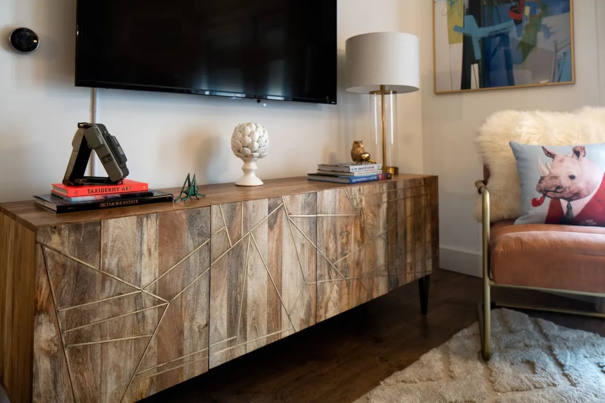 A cozy and stylish living area with a wooden sideboard featuring geometric patterns, a wall-mounted television, a decorative lamp, an artistic pig-themed pillow, and a chic armchair. The space is inviting and set up for relaxation or informal conversation.