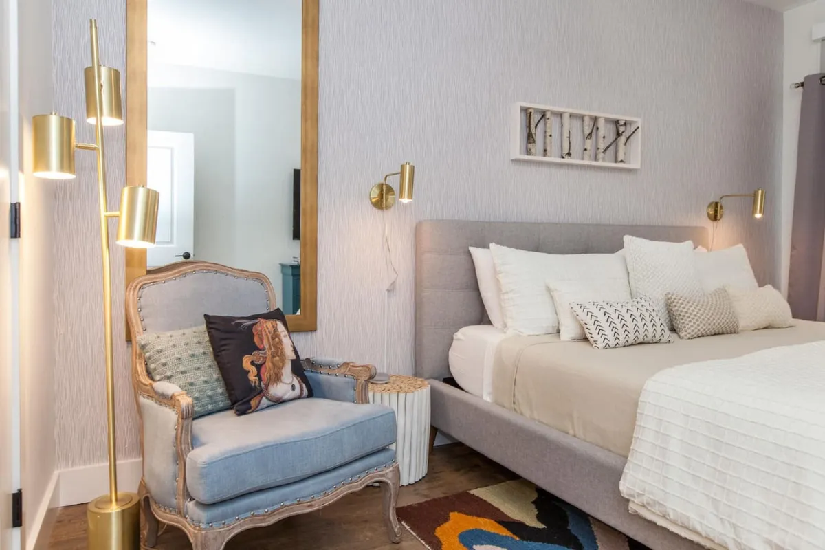 A cozy bedroom with a bed dressed in decorative pillows, a vintage-style chair next to it, a gold floor lamp, and a decorative mirror—all bathed in inviting, warm light. The setup radiates intimacy and comfort.