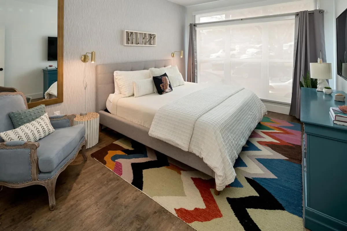 A bright, stylish bedroom featuring a plush bed adorned with decorative pillows, a colorful area rug beneath, an inviting armchair, and a large window letting in ample natural light. The setup evokes comfort and tranquility, perfect for relaxation.